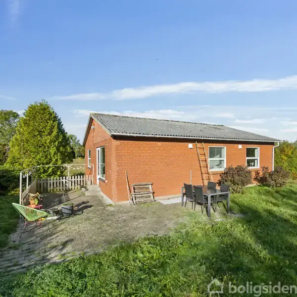 Et rødt murstenshus står stille i en grøn have. Der er en terrasse med havemøbler og en trillebør foran en blå himmel.