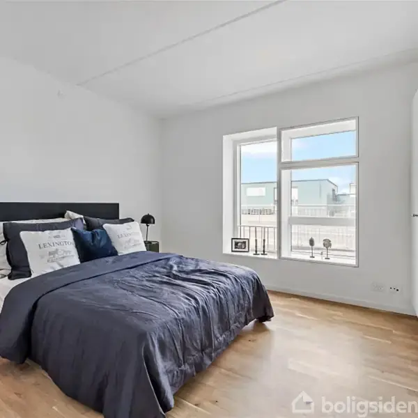 Soveværelse med en dobbeltseng dækket af mørkt sengetøj, flankeret af natborde med lamper. Et stort vindue bader rummet i naturligt lys, med en indbygget skabsplads til højre.