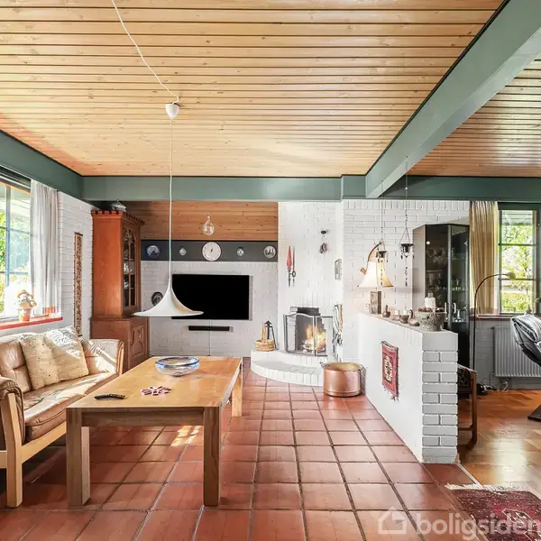 Stue med træloft har et sofaarrangement og træbord. Pejs og TV er placeret langs en væg. Store vinduer lukker lys ind, og der er planter og pyntegenstande omkring.