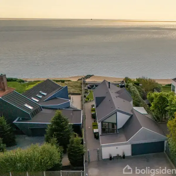 Moderne huse ligger langs en kystlinje; de er omgivet af grønne træer med en udsigt over havet i baggrunden.