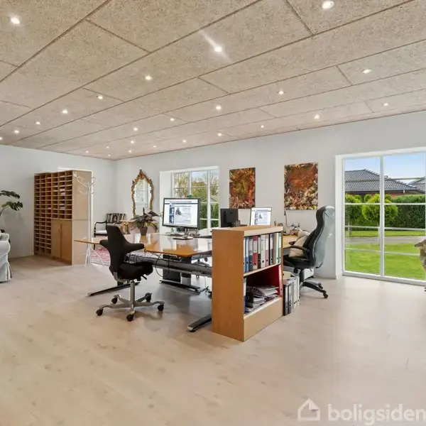 Et rummeligt kontor er indrettet med store skriveborde, kontorstole og computere. Vinduet i baggrunden giver udsigt til en grøn have. Væggene prydes af kunstværker.