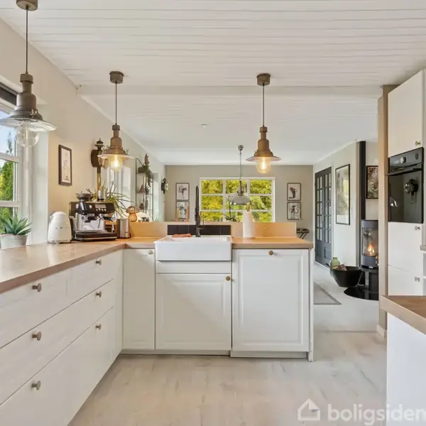 Et lyst køkken med hvide skabe og træbordplader, indeholder en central køkkenø med vask. Rummet er oplyst af hængende lamper og åbner op til en lys stue.