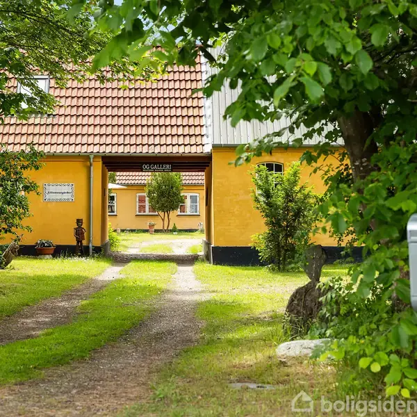 Gul bygning med rødt tegltag omkranser en gårdsplads med græsareal. En grusvej fører ind mod gården. Et skilt på bygningen viser teksten "O.G Galleri". Postkasse med tallet "5" ses til højre.