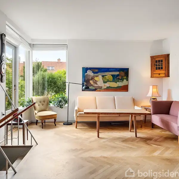 Stuen har møbler som en sofa, lænestol, skrivebord og flere kunstmalerier på væggene. Store vinduer giver udsigt til en have. Elegant trægulv og moderne indretning.