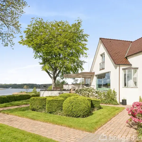 Et hvidt hus med rødt tag står ved en sø. Huset har store vinduer og er omgivet af en velplejet have med blomster og buske.