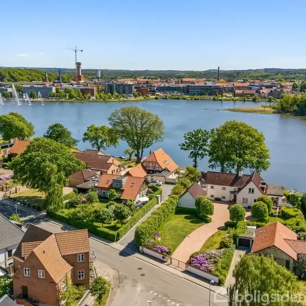 Boligerne ligger i et grønt villakvarter ved en sø. Trækroner og velholdte haver omkranser det stille nabolag. I baggrunden ses en by med bygninger og en kran.