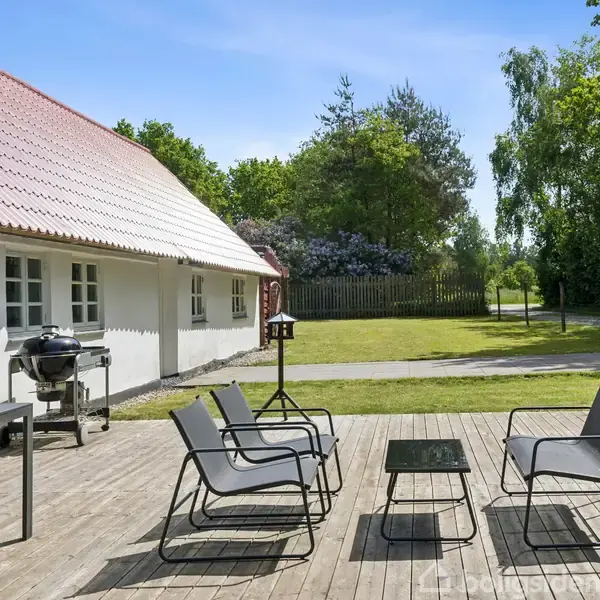 Hvide stole og et lille bord står på en træterrasse. Ved siden af er en grill. Huset i baggrunden er hvidt med rødt tag. Haven er omkranset af træer.