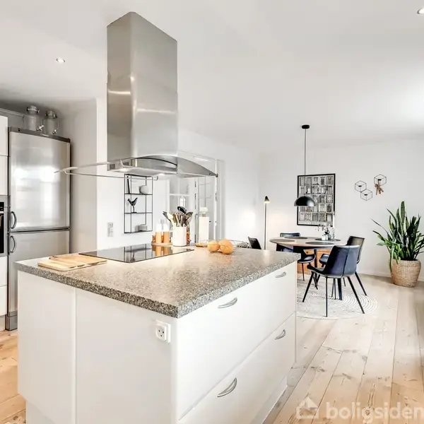 Et hvidt køkken med en stor kogeø, hvor der står frugt og en knivblok. I baggrunden ses et spisebord med stole og en plante i et lyst rum.