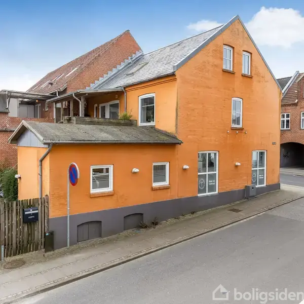 En orangerød murstensbygning står på et hjørne af en byvej, omgivet af andre røde murstenshuse. Skiltet ved siden af huset angiver parkeringsforbud.