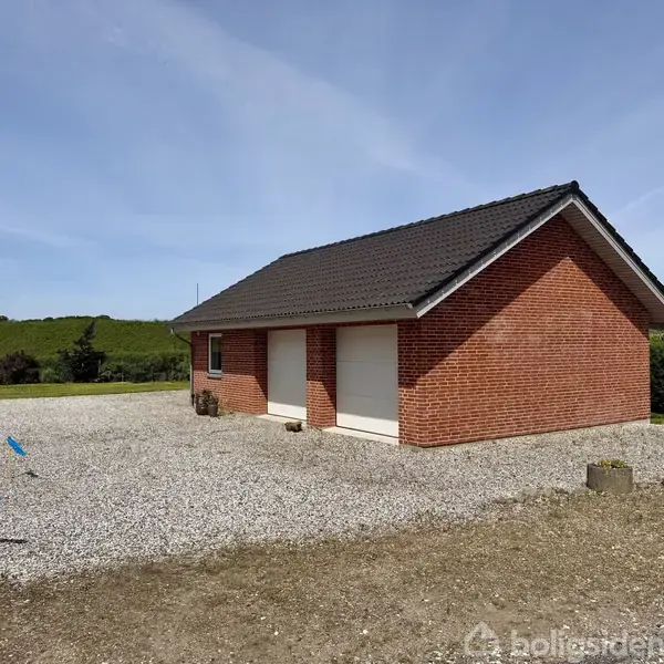 Rødt murstenshus med sort tag står på en grusbelagt gårdsplads. To lukkede hvide garageporte er synlige. Haven har græs og buske i baggrunden, klar blå himmel over.