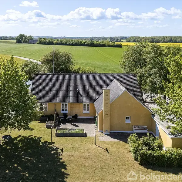 Et gult hus står omgivet af grønne træer og marker under en blå himmel med skyer. Tætliggende buske og en vej ses i baggrunden.