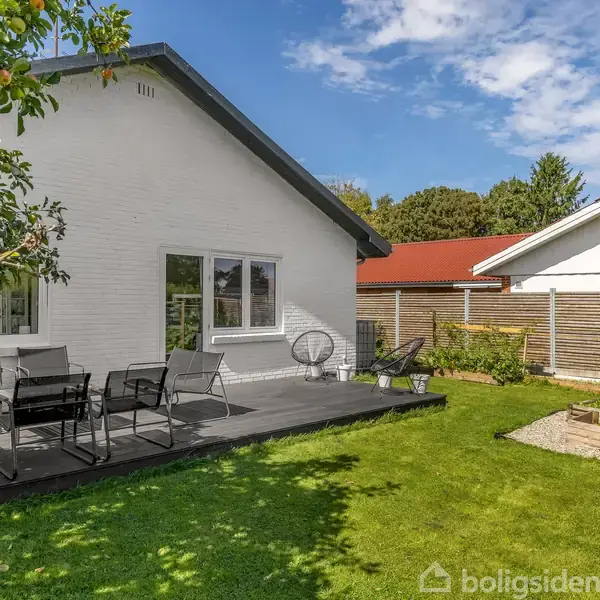 Hvidt hus med terrasse, hvor der står sorte havemøbler. Terrassen er omgivet af en grøn græsplæne med højbede og træhegn i baggrunden. Rød tagryg på nabohuset.