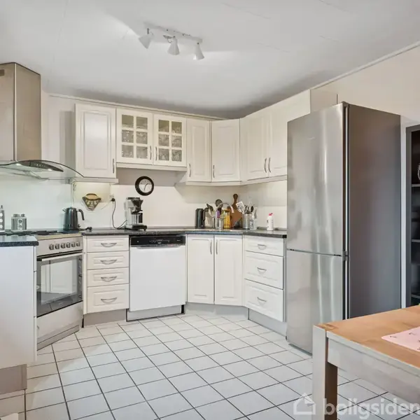 Køkken med hvide skabe og apparater som komfur, emhætte, og opvaskemaskine. Der står en høj, sølvfarvet køleskab ved siden af en hylde fyldt med køkkenting. Bord i forgrunden.