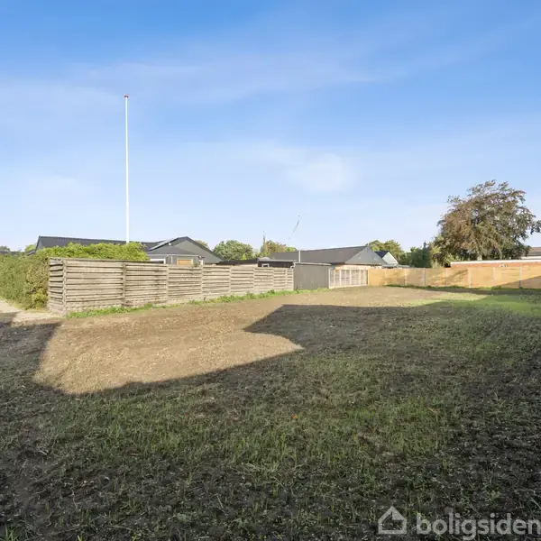 En tom grund med jævn jord under en blå himmel omgivet af huse og hegn. Ingen tekst er synlig i billedet.