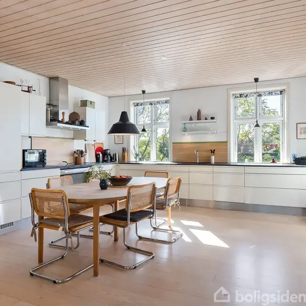 Et lyst, moderne køkken med hvide skabe og træloft. Et spisebord med fire stole står i midten, og vinduer lader naturligt lys strømme ind. En lampe hænger over bordet.