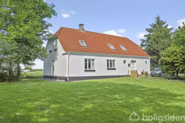 Et hvidt hus med et rødt tegltag står omgivet af en grøn have med træer og en bil parkeret ved siden af.