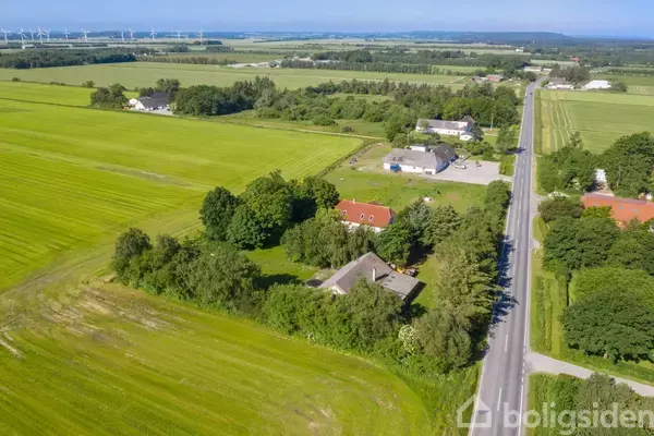 Landejendom omgivet af træer og marker ligger langs en lang, asfalteret vej i en landlig region med vindmøller i baggrunden.