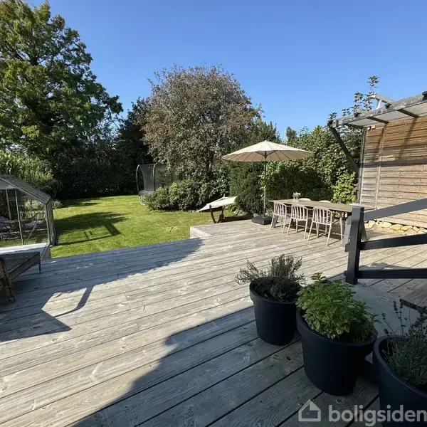 En træterrasse med potteplanter står i sollys. Ved siden af er et spisebord og parasol, mens en have med græsplæne og drivhus ses i baggrunden.