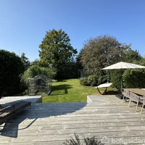 Terrasse med træplanker ligger foran en have med græsplæne og buske. Til højre er et træbord med stole og parasol. Et drivhus står på græsset under en blå himmel.