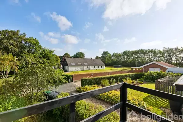 En træterrasse med en sort rækværk giver udsigt til en stor gård med hvid bygning i landlig omgivelser, inkluderende grønne marker, træer og en rød udhusbygning.