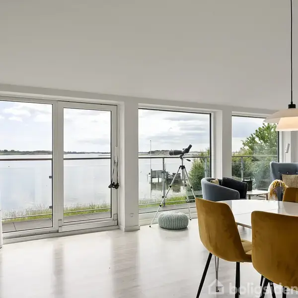Spisestue med hvide vægge og gulv, runde spisebord omgivet af gule stole. Store vinduer med udsigt over vand og teleskop ved siden af. Lænestol i hjørnet.