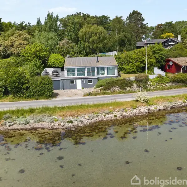 Hus beliggende tæt på vandet, omgivet af grønne træer og buske. Der er en vej mellem huset og vandkanten. Bil parkeret ved siden af huset.