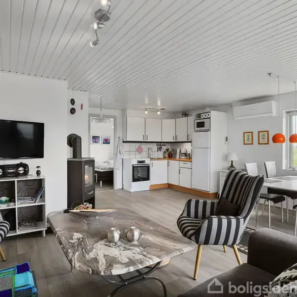 Stue med stribede stole og sofabord i forgrunden; køkken i baggrunden med spisebord ved vindue. Fjernsyn på væggen, loftsbelysning, og trægulv. Lys og moderne indretning.