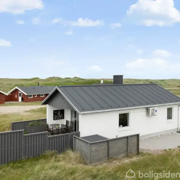 Hvidt hus med sort tag står stille omgivet af fladt græsareal. Terrasse med hegn ses ved huset, og i baggrunden er der flere huse. Himlen er delvist skyet.
