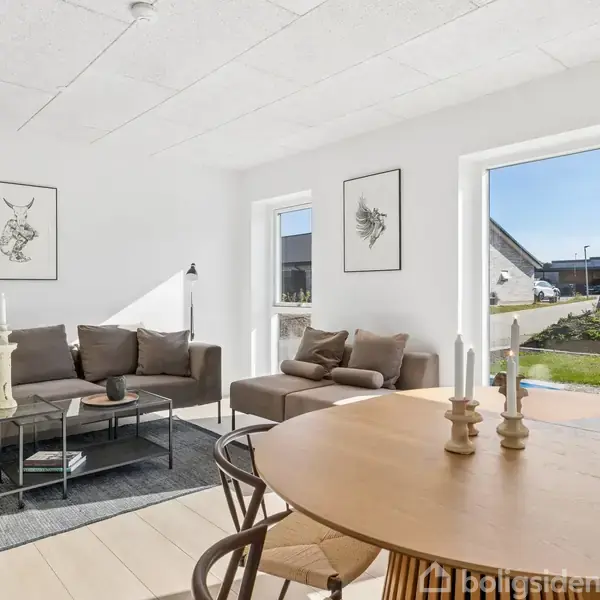 En stue med en grå sofa og sofabord står ved siden af vinduer, der giver udsigt til en have. Et spisebord med stole står i forgrunden. Væggene er prydet med billeder.