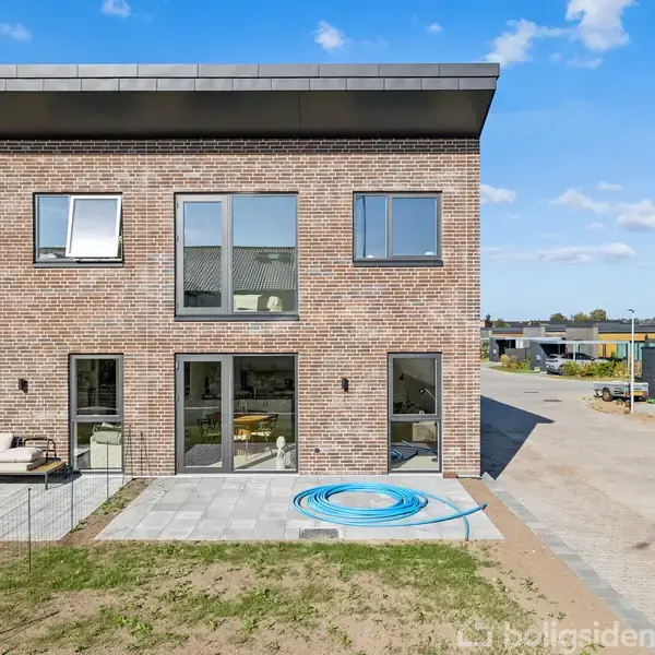 To-etagers murstenshus med store vinduer og terrasse. En haveslange er rullet op foran huset. I baggrunden ses en vej og et andet hus under blå himmel.
