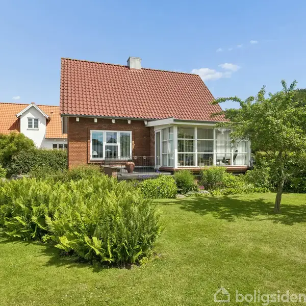Hus med rødt tegltag og stort vinduesparti står i en velplejet have med grønne buske og træer. En blå himmel danner baggrund.