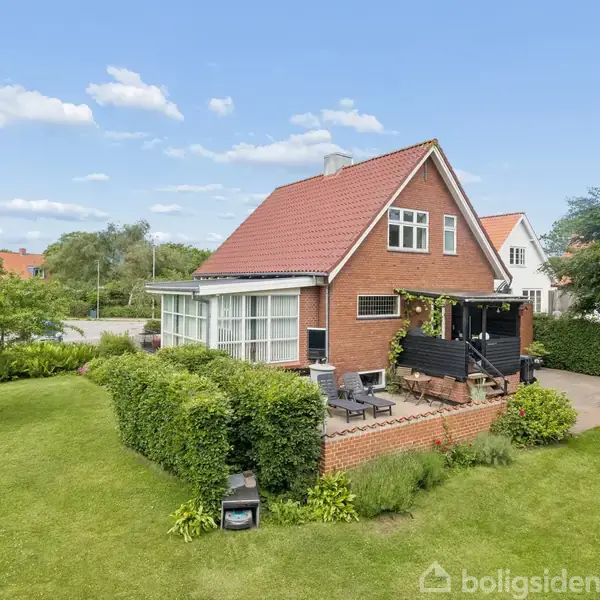 Rødt murstenshus med rødt tag står i en grøn have med terrasse og havemøbler. Omkranset af buske og træer under en blå himmel med spredte skyer.