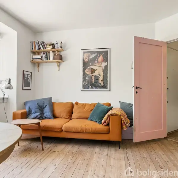 Orange sofa med farverige puder står foran en hvid væg med kunstplakater. Ved siden af er en lyserød dør åben ind til et rum med jakker. Trægulv og vindue mod grøn udsigt.
