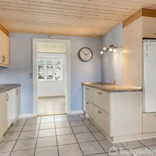 Køkken med lyst inventar og flisegulv. Et køleskab står mod højre, mens en vask er mod venstre. Et ur hænger på væggen, lys strømmer ind fra en dør.