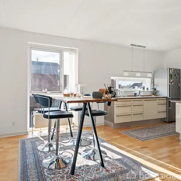 Rundt spisebord med barstole står i et lyst køkkenområde. En stor rektangulær lampe hænger over køkkenøen, med køleskab og skabe i baggrunden. Tæpper ligger på trægulvet.