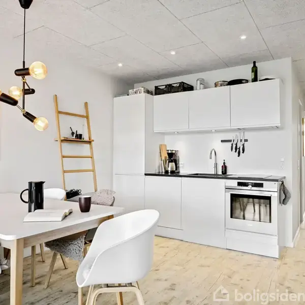 Terrassemøbler står på en flisebelagt gårdsplads foran et moderne hus med store vinduer. Et sort bord og stole er centreret med potteplanter og omgivende skov i baggrunden.