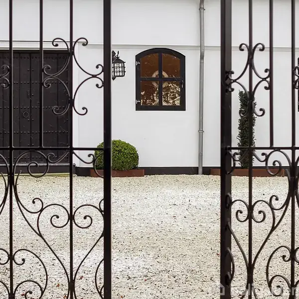 Dekorativ smedejernsport står åben mod en hvid bygning med sorte porte og vinduer. I gården er der grus og arrangerede planter i potter ved væggen.