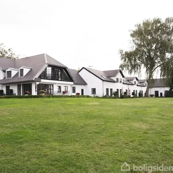 En stor, moderne hvid bygning med sort tag står på en velplejet græsplæne. Der er store vinduer og træer rundt om.