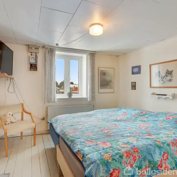 Soveværelse med en stor seng dækket af et blomstret tæppe. En stol står ved siden af en væghængt TV. Vindue med gardiner i midten af væggen. Væggene prydet med billeder.