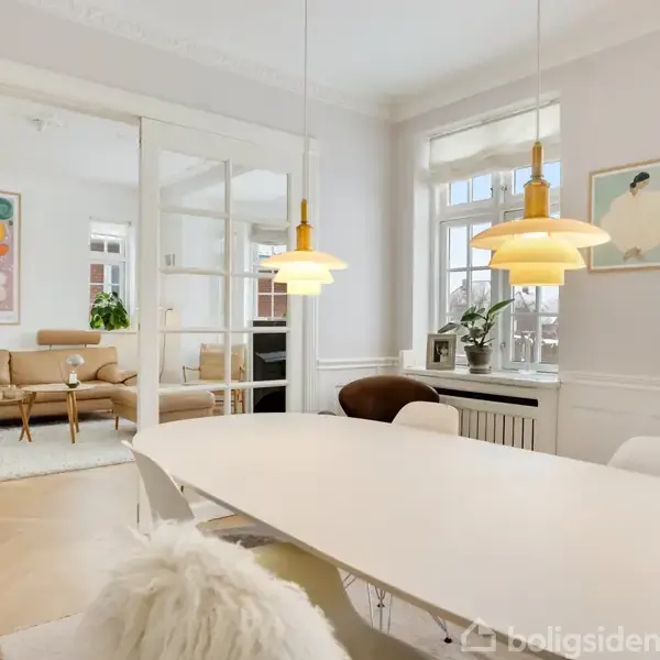 Et spisebord med stole står i et lyst rum. To hængelamper lyser bordet op. I baggrunden ses en stue med sofa og farverig kunst på væggen.