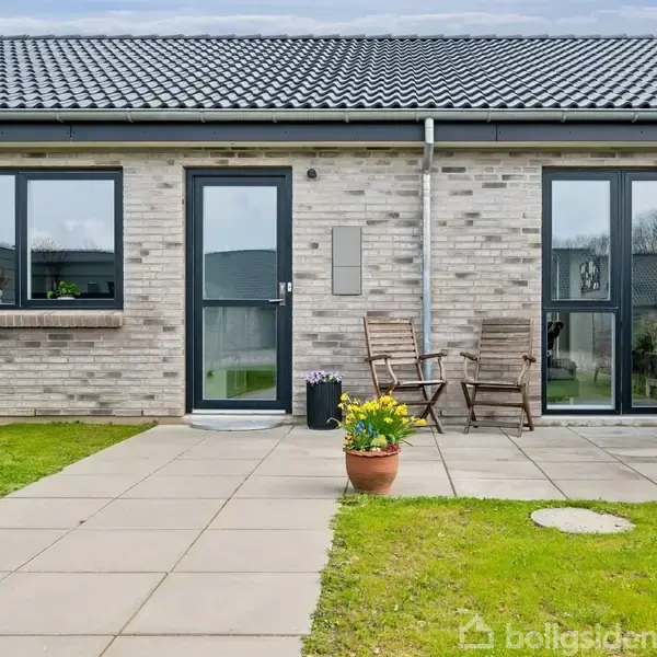 En murstensbygning med to store vinduer og en dør. To stole og en potte med blomster står på en flisebelagt terrasse i en græsplæne i boligområde.