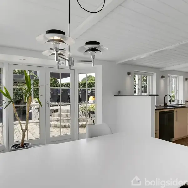 Lyse køkkenområde med hvidt bord og moderne lamper. Store glasdøre fører ud til en terrasse med planter. Køkkenet har lyse træskabe og hvidmalet loft.