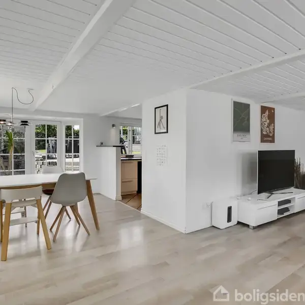 Spisestue med træbord og stole står foran vinduer, der lader naturligt lys ind. Stuen har en sofa, TV og lyse trægulve, omgivet af hvide vægge og loft.