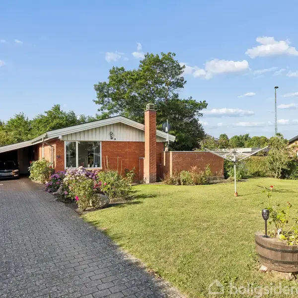 Rødt murstenshus med carport ligger i en grøn have. En flisebelagt indkørsel fører til huset, omgivet af blomsterbede. En grill og tønde står på græsplænen under en blå himmel.