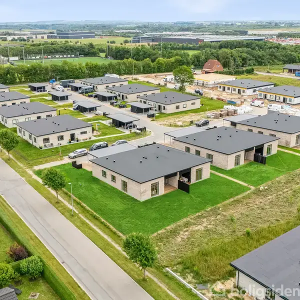 Flere ens-formede enfamiliehuse med flade tage er placeret i rækker i en nyudviklet forstad. Veje og grønne områder omgiver husene, som ligger i et åbent landskab.