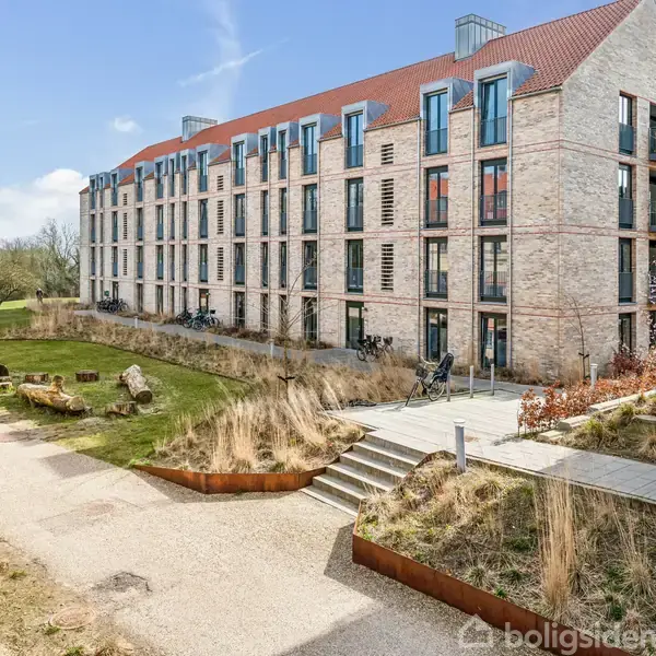 Bygning med rødt tegltag og store vinduer, omgivet af græs og stier, cykler står op ad muren, og der ses en havebænk langs stien.