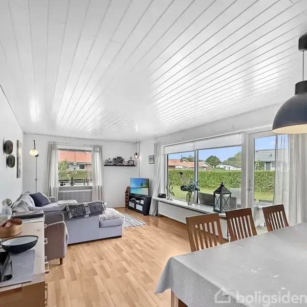 Stue med sofa og TV står ved et vindue med udsigt til en have. En spiseplads med bord og stole er til højre, omgivet af hvide vægge og træloft.
