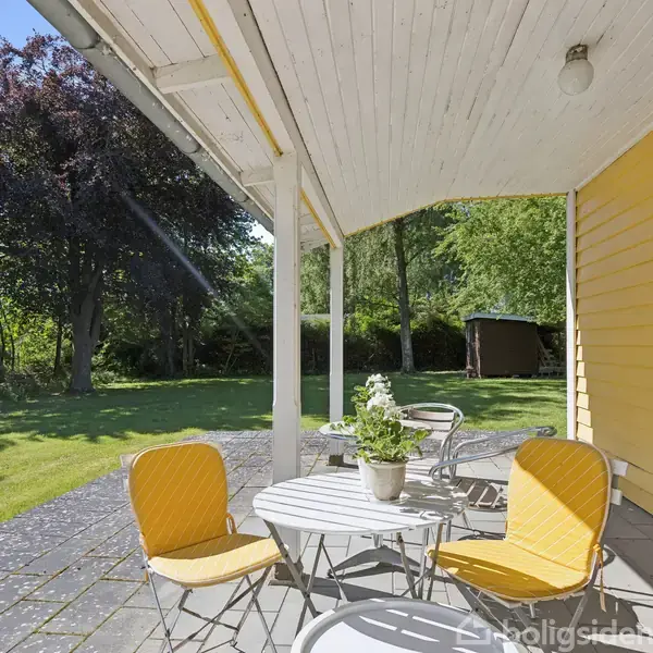 Terrassemøbler med gule stole står på flisebelagt terrasse ved gul bygning med overdækket tag. En plante i potte står på bordet, omgivet af en grøn have med træer og skur.