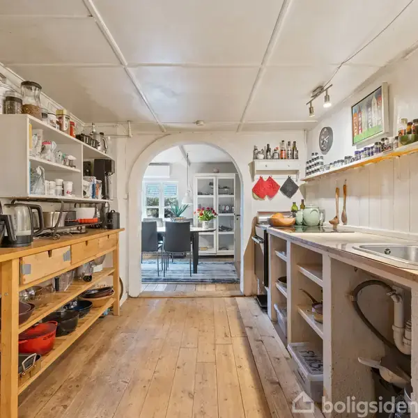 Et køkken med åbne hylder fuldt af køkkenredskaber, en træbænk med apparater og en vask. Gennem buet døråbning skimtes et spiseområde. Veggene er lyse, og gulvet er af træ.