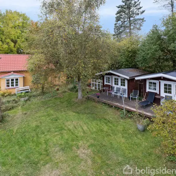 To huse står i en frodig have; det ene er gult med rødt tag, det andet brunt med terrasse og havemøbler, omgivet af træer og buske.